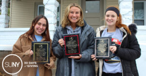 Three female SUNY Cortland students stand outside their apartment in winter jackets holding plaques of acknowledgement from the city fire department.
