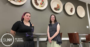 Two female SUNY Oswego students stand in front of the wall art display titled Potluck, showing plates of food made out of various fabrics.