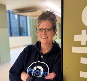 Shelly Dunham leans against a green wall with text while wearing an OCC sweatshirt