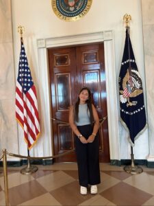  Annalisa Alicea stands in front of a wooden door. The door is flanked by a U.S. flag & a flag with a U.S. seal.