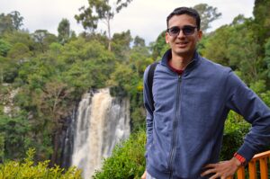  Kaustubh Sathe stands in a blue jacket with a waterfall and a forest environment behind him.