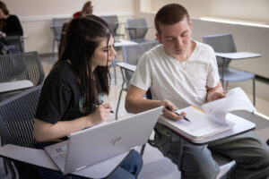 Two students sit at desks and look over a notebook while one has an open laptop.