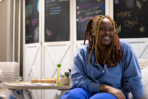 A student sits and smiles next to a smll table. A wall with chalkboards and writing sits behind them.