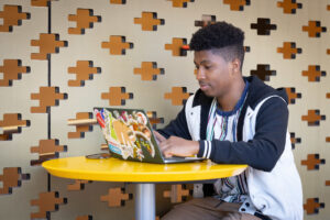 A student sits at a small table and works on a laptop covered in various stickers