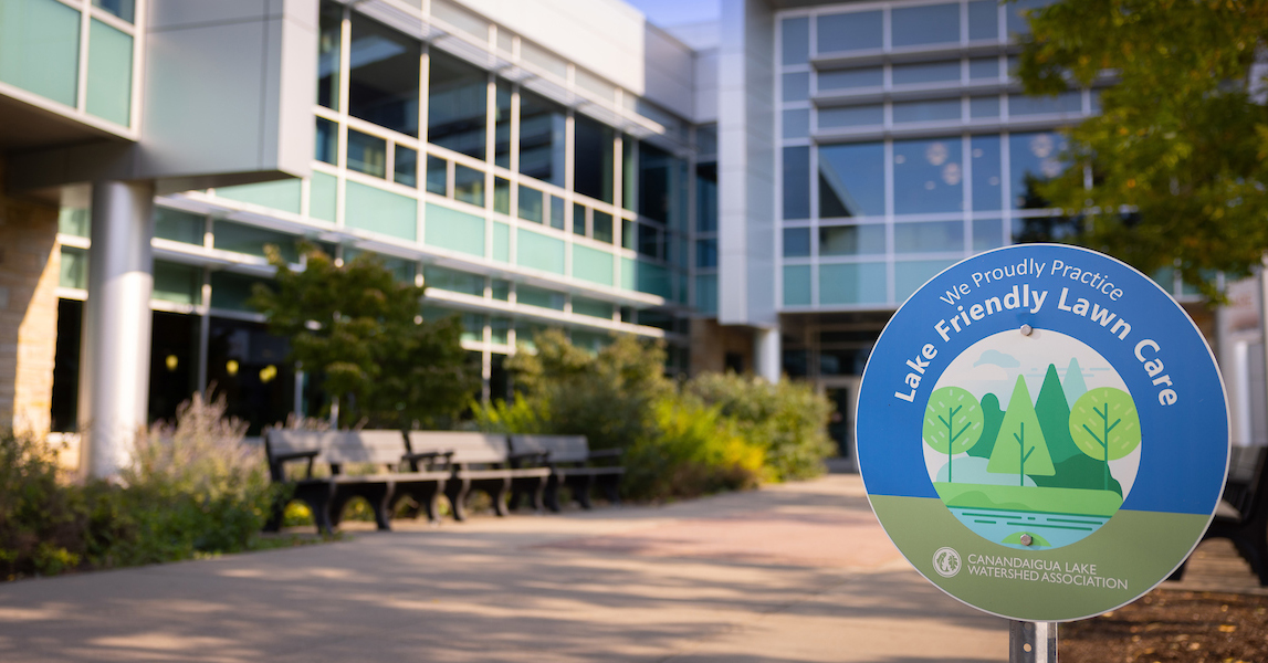 A sign that reads "we practice lake-friendly lawn care" in front of a large steel, concrete, and glass building at Finger Lakes Community College