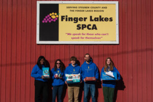 Corning Community College students pose with various items outside of a red building with a sign that reads Finger Lakes SPCA