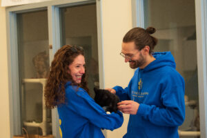 Two students smile as one holds a black cat and the other goes to pet the cat