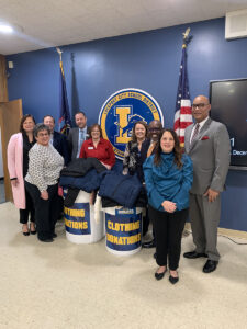 SUNY Niagara staff pose with a donation of winter clothing in front a wall featuring the Lockport School District logo.