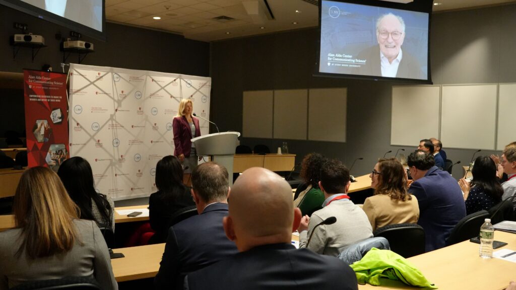 A presenter stands at a podium in a lecture room delivering remarks to an audience seated at tables. A large screen above displays a video image of an older speaker with the text “Alan Alda Center for Communicating Science at Stony Brook University.” Audience members face the presenter, with microphones, notebooks, and water bottles visible on the tables, and a branded backdrop and banner appear behind the podium.