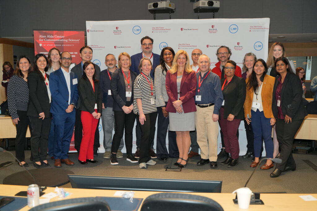 A large group of participants stands together at the front of a conference room posing for a group photo. Behind the group is a backdrop featuring SUNY and Alan Alda Center for Communicating Science logos, along with a branded banner promoting science communication. Tables and seating are visible in the foreground.