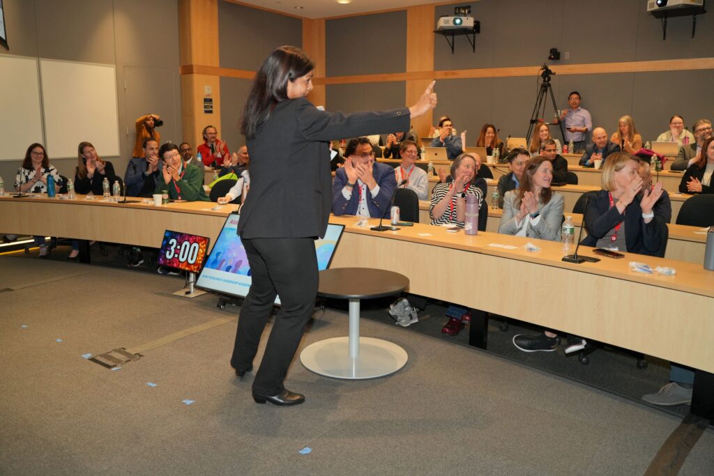 A presenter stands at the front of a classroom facing an audience seated at tiered tables. The presenter gestures toward the audience while a digital timer on a screen nearby reads “3:00.” The audience appears attentive and several individuals are clapping, with water bottles, notebooks, and laptops on the desks.