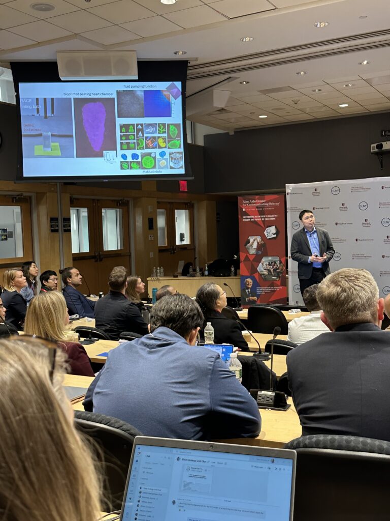 A presenter stands in front of a classroom audience delivering a talk while gesturing toward a large screen. The slide shows images and diagrams related to scientific research, including bioprinted materials and labeled visuals. Attendees sit at tables with laptops, notebooks, and water bottles, facing the speaker.