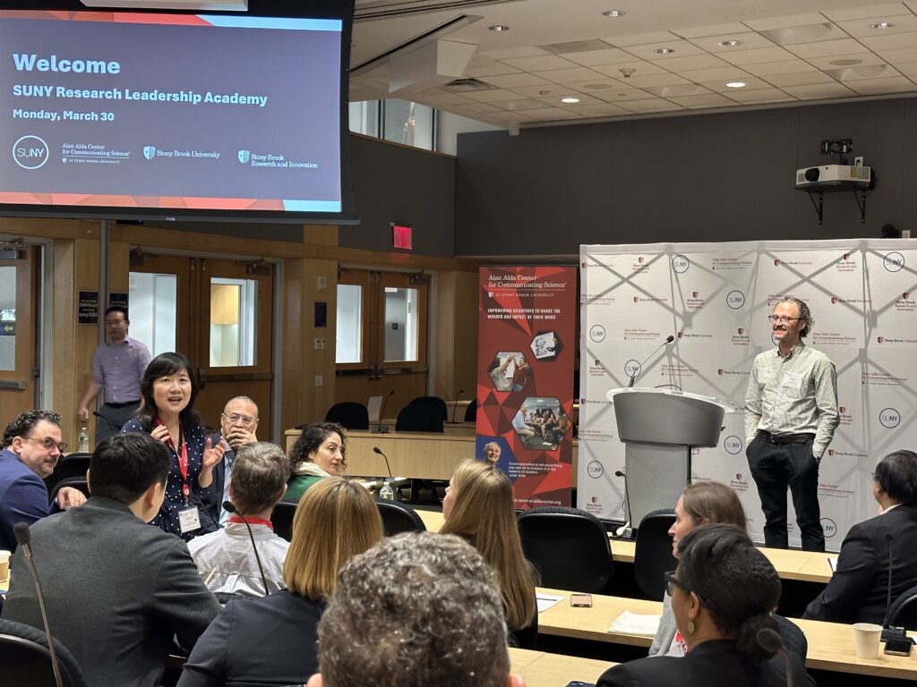 A lecture room with attendees seated at tables facing a presenter at the front. A large screen displays a welcome slide reading “SUNY Research Leadership Academy, Monday, March 30,” alongside SUNY and Alan Alda Center for Communicating Science logos. One audience member stands and speaks while the presenter stands near a podium listening.