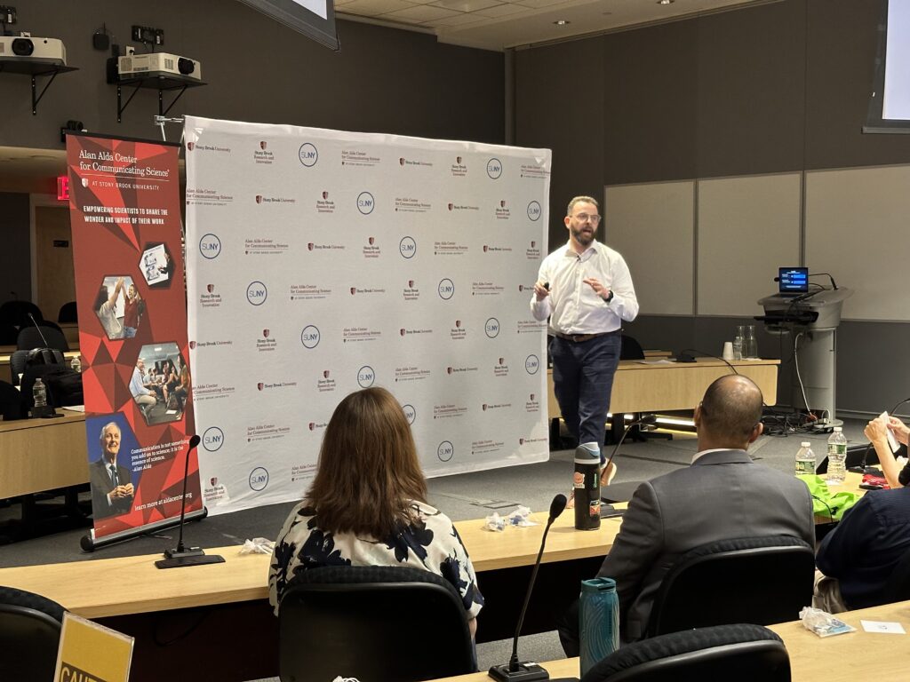 A presenter stands at the front of a classroom speaking to an audience seated at tables. Behind the presenter is a backdrop with SUNY and Alan Alda Center for Communicating Science logos. A vertical banner reads “Alan Alda Center for Communicating Science” with the tagline “Empowering scientists to share the wonder and impact of their work.”