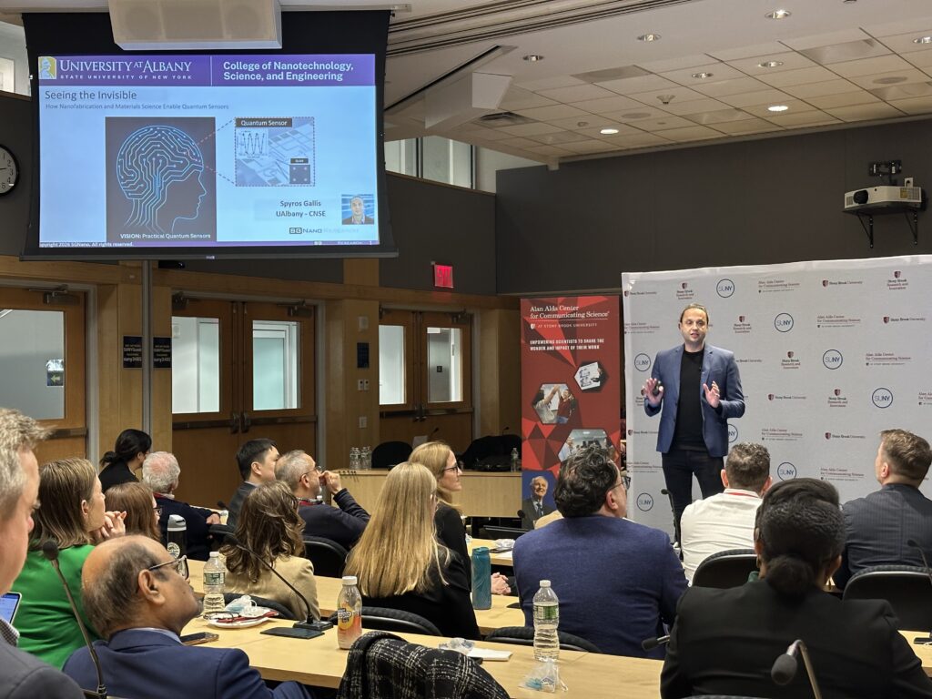 A speaker stands at the front of a lecture room presenting to an audience seated in rows. A large screen displays a slide titled “Seeing the Invisible” from the University at Albany College of Nanotechnology, Science, and Engineering, including a graphic of a human head with circuit lines and references to quantum sensors. An event banner for the Alan Alda Center for Communicating Science stands nearby.