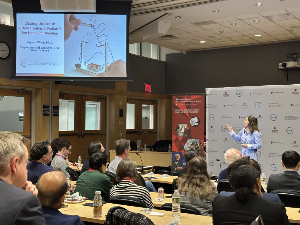 A presenter stands at the front of a lecture room addressing an audience seated in rows. A large screen displays a slide titled “Closing the Loop: A New Frontier in Pediatric Eye-Hand Coordination,” credited to Jingyun Wang, Ph.D., from the SUNY College of Optometry. The slide includes an image of a hand guiding a wire loop through a shaped metal path.